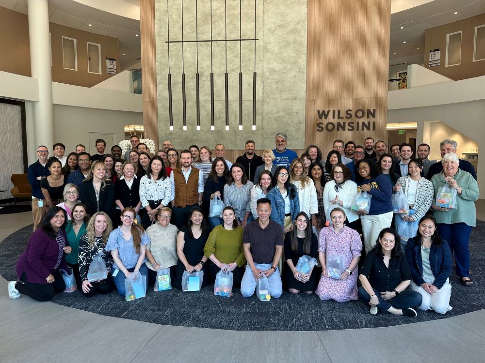 Group photograph shows roughly 50 people gathered in a Wilson Sonsini lobby, Participants pose in several rows with smiles, gift bags, and Wilson Sonsini signage visible behind them.