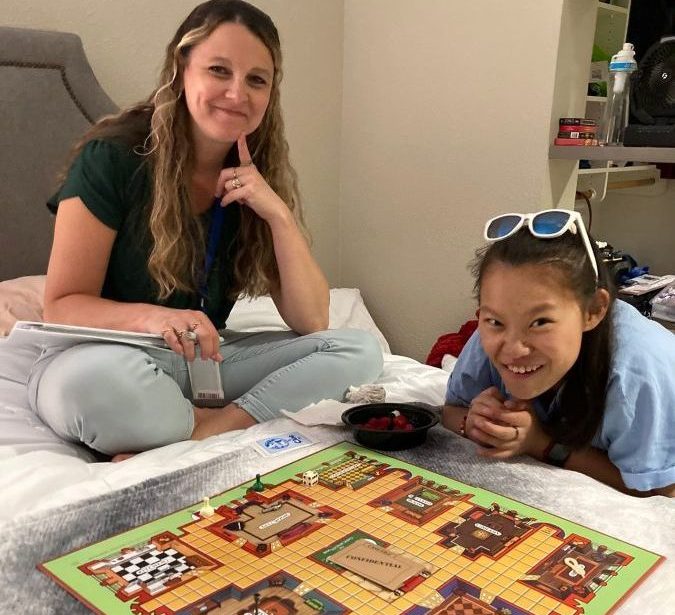 Photograph shows two women smiling while playing a colorful mystery-themed board game on a bed. Game board features rooms, pathways, character pieces, dice, cards, and snack bowl, creating a casual indoor recreation scene.