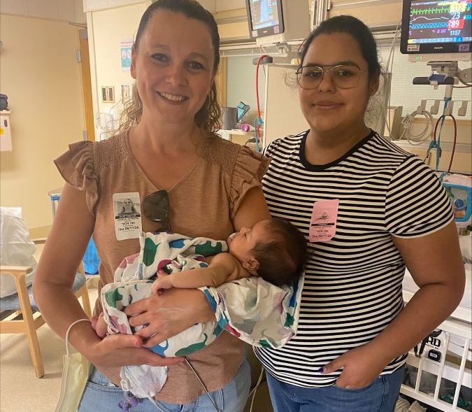 Photograph of two women smiling in a hospital room while holding a newborn wrapped in a patterned blanket. Medical monitors, equipment, hospital beds, and identification badges indicate a maternity or neonatal care setting.