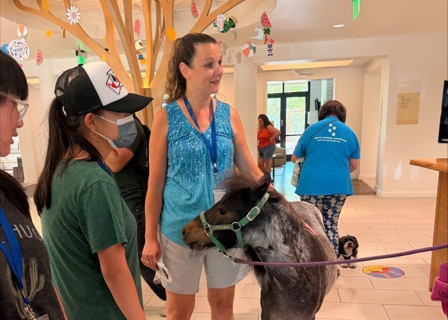 Photograph shows an indoor animal-assisted activity, with a woman guiding a miniature donkey through a decorated community facility. Children and adults gather nearby, while colorful hanging crafts, a window, and a purple barrier frame scene.