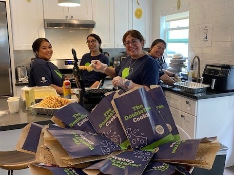 Photograph shows four smiling staff members preparing food in a bright kitchen. Foreground features stacked purple Double Good popcorn bags and waffle trays, while stainless appliances, countertops, and serving supplies fora food-service event.