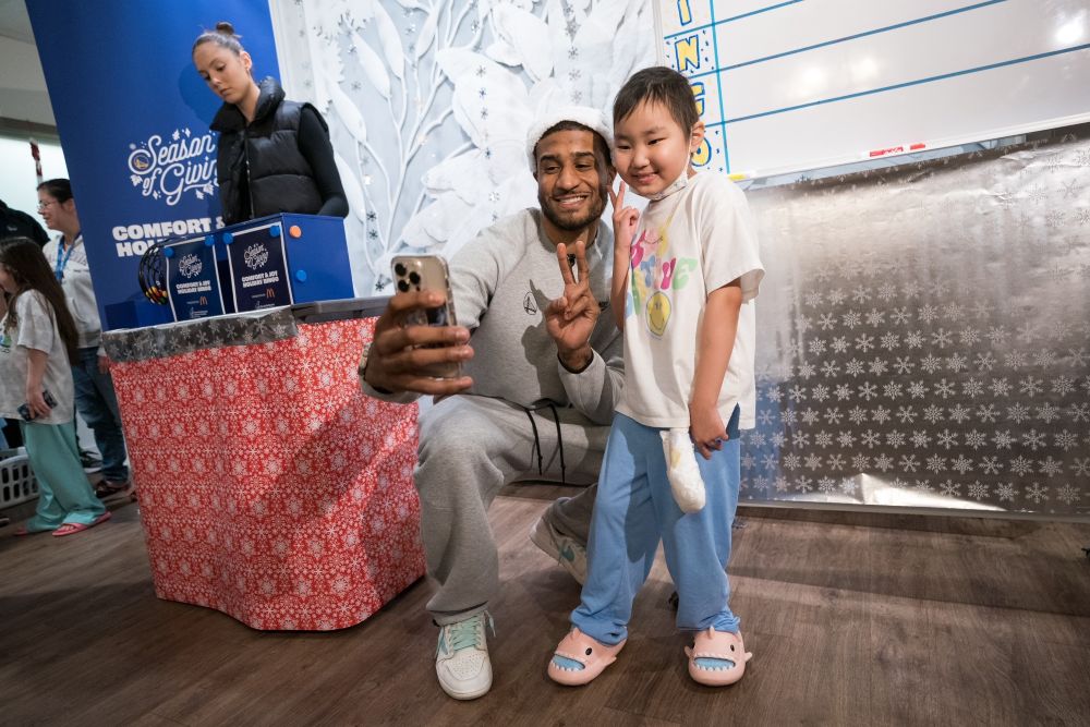 Photograph shows a GP2 and a child taking a selfie at a holiday event beside a large red gift-wrapped table. Snowflake decorations, a branded “Season of Giving” display, and a whiteboard form a festive backdrop.