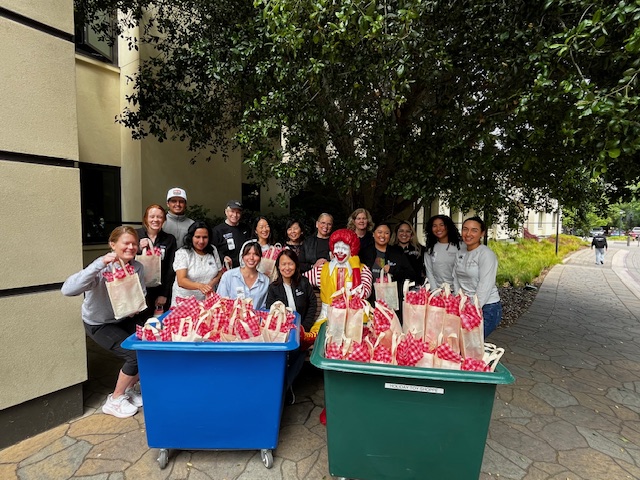 Photograph shows a group of volunteers gathered outside a building for a community care kit event. Red-and-white packaged treats fill blue and green rolling bins.