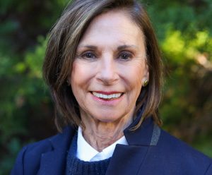 Linda Howell: Woman wearing a collar shirt and sweater smiling. There is a greenery behind.