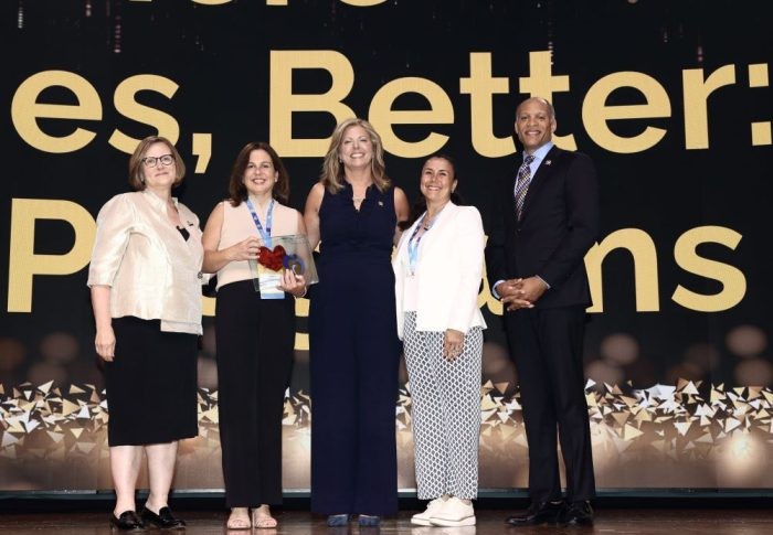 Five people standing on stage smiling as an RMHC Bay Area team member holds a heart-shaped award in front of a gold-themed presentation screen.