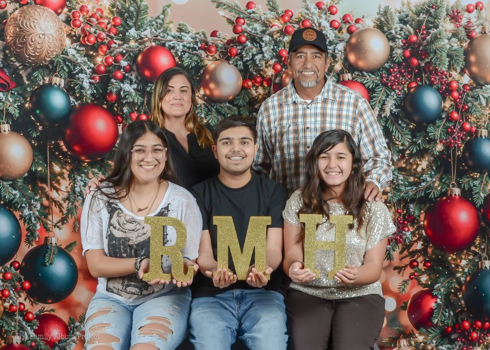 The Medina Flores family posing in front of a festive holiday backdrop holding gold RMH letters.