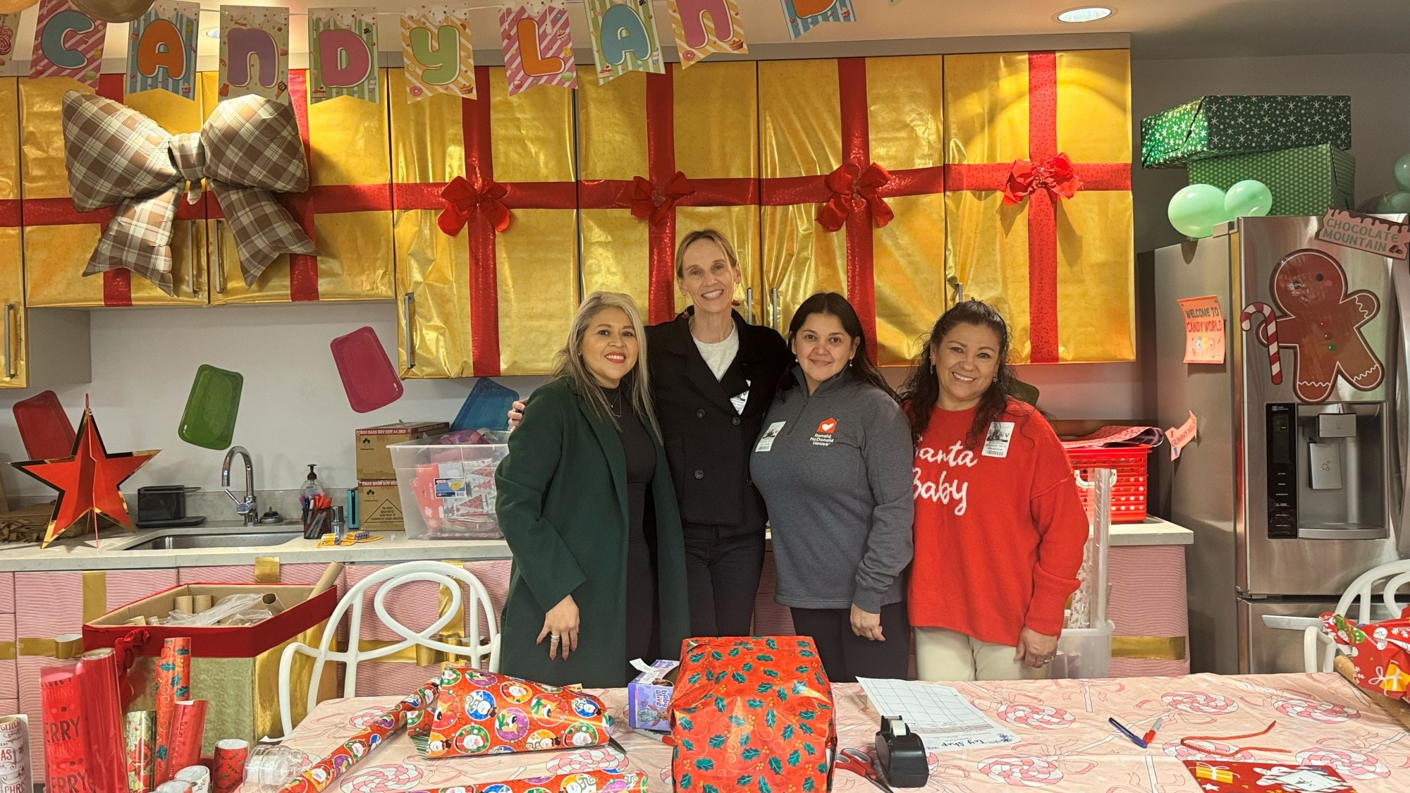 Photograph shows four women posing behind a festive candy-themed gift-wrapping table. Colorful presents, wrapping paper, oversized bow, balloons, holiday decorations, and candy-shop displays create a cheerful seasonal setting.