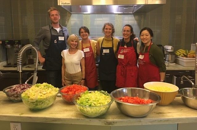 Group of smiling volunteers standing behind a counter filled with bowls of chopped vegetables in a kitchen.