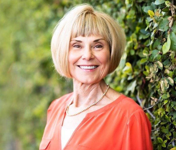Sally Duplantier: Smiling woman with a blonde bob haircut wearing an orange blouse and pearl necklace, standing in front of a leafy green wall.